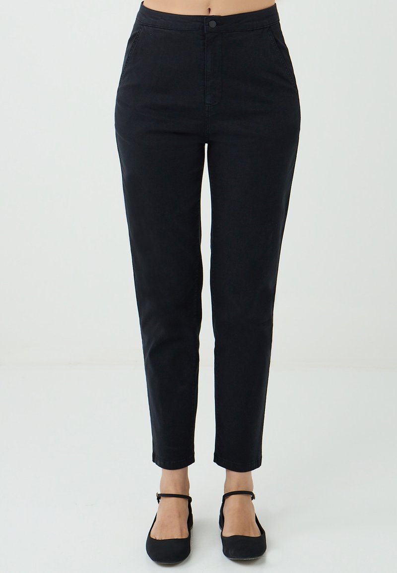 Pantalons noirs taille haute associés à des ballerines noires, dotés d'une simple fermeture à bouton et à zip, présentés sur un fond neutre.