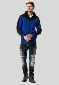 Blaue und schwarze Zip-Hoodie mit Kapuze, ausgestattet mit einem Chevron-Design und Fronttaschen. Kombiniert mit einer multi-pocket, distressed Denim-Jeans.
