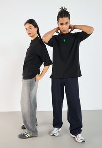 Svarte t-skjorter med en grønn diamantlogo, kombinert med løse grå og mørkeblå jeans. Personene har på seg sneakers og står mot en enkel bakgrunn.