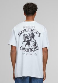 Hvid bomulds T-shirt med et grafisk design af to løver, der kæmper på ryggen, med teksten: "FARLIGE JORDER" og "OVERLEVELSE FOR DE STÆRKESTE."