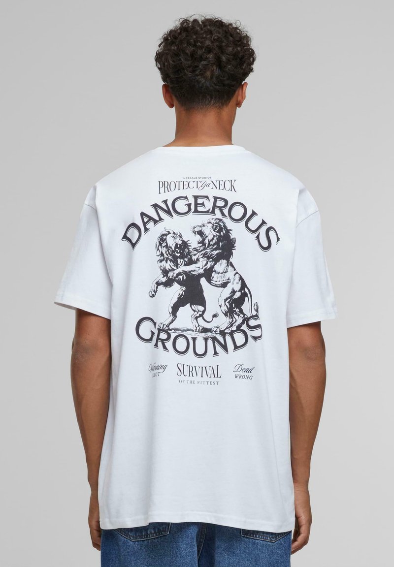 Hvid bomulds T-shirt med et grafisk design af to løver, der kæmper på ryggen, med teksten: "FARLIGE JORDER" og "OVERLEVELSE FOR DE STÆRKESTE."