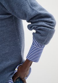 Bras portant une manche de pull bleu en peluche remontée, révélant un poignet de chemise rayé bleu et blanc et une main dans une poche.