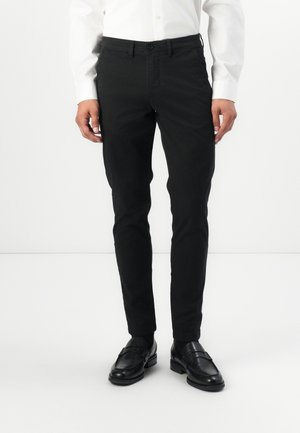 Pantalones slim fit negros fabricados en una mezcla de algodón, con una textura suave y bolsillos frontales, llevados con mocasines negros y una camisa blanca.