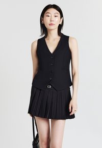 Eine Frau trägt eine ärmellose schwarze Knopfveste und einen gefältelten schwarzen Minirock und hält eine schwarze Handtasche vor einem schlichten Hintergrund.