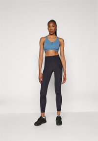 Ανοιχτό μπλε αθλητικό σουτιέν με κουμπιά, συνδυασμένο με ψηλόμεσα μαύρα κολάν. Το μοντέλο φοράει μαύρα αθλητικά παπούτσια. Απλός, εφαρμοστός σχεδιασμός.