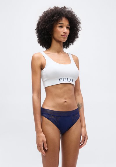 Γυναίκα με σγουρά μαλλιά που φοράει λευκό κοντό τοπ με την επιγραφή "POLO" και μπλε σλιπ από δαντέλα, στέκεται μπροστά σε απλό φόντο.