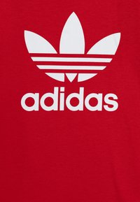 Raudona medvilninė marškinė su didele balta Adidas logotipu, kuris turi trijų lapų dizainą, o po užrašu yra trys horizontali juostelės.