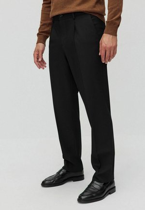 Zwarte broek gemaakt van een gladde stof, met een rechte pasvorm, plooien, zijzakken en een tailleband met ri loops. Gecombineerd met zwarte instappers.