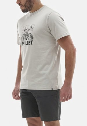 Homme portant un t-shirt gris clair avec un motif de montagne et le texte "MILLET", associé à un short noir, debout devant un fond blanc.