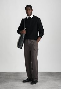 Μαύρος ριγέ πουλόβερ, λευκό πουκάμισο με γιακά, γκρι παντελόνι cargo, μαύρα δερμάτινα παπούτσια και γκρι τσάντα ώμου. Απλό φόντο.