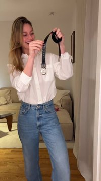 Sort bælte med præget mønster og sølvspænde, båret af en person i en hvid langærmet skjorte og blå jeans. Neutral baggrund.