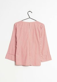 Dlouhá blůza s vertikálními červenými a bílými pruhy a kulatým výstřihem, vystavená na černém ramínku proti bílému pozadí.