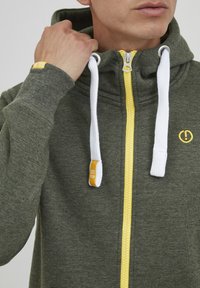 Groene hoodie met ritssluiting en gele rits, witte trekkoorden en een logo. Gemaakt van een katoenmix met een zachte textuur en ribbelmanchetten.