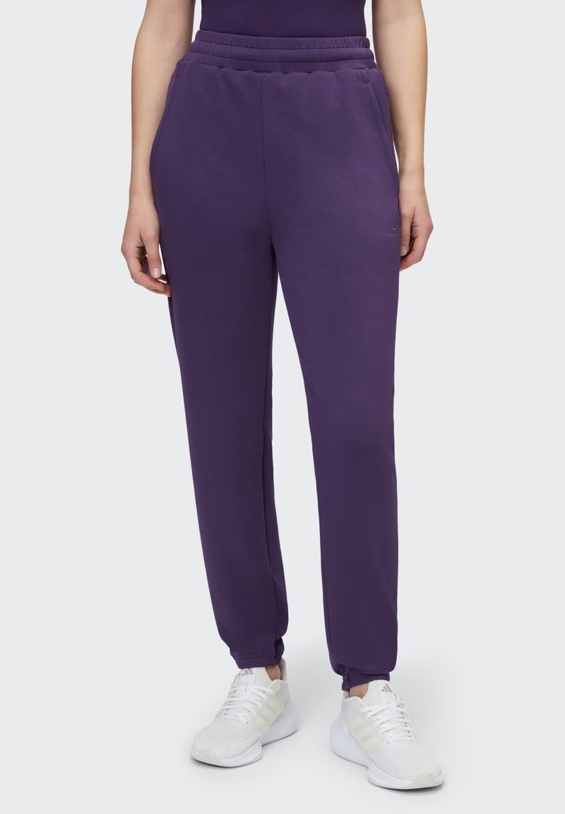 Lila Jogginghose mit elastischem Bund und Bündchen an den Knöcheln, mit glatter Textur und Seitentaschen, kombiniert mit weißen Sportschuhen.