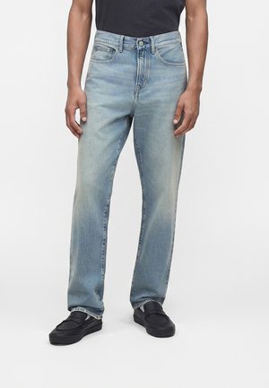 Homme portant un jean droit bleu clair, des chaussures noires sans lacets et une chemise noire visible à partir du milieu du torse, debout devant un fond blanc.