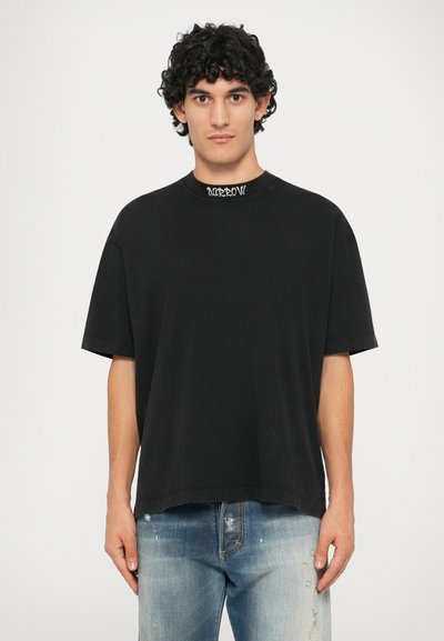 Svart oversized t-skjorte med rund hals, med en teksturert trykt etikett på kragen og korte ermer. Kombinert med falmede blåjeans.