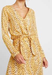 Kvinna som bär en långärmad klänning med gul leopardmönster, framknäppning och knytbart midjebälte, stående mot en enhetlig bakgrund.