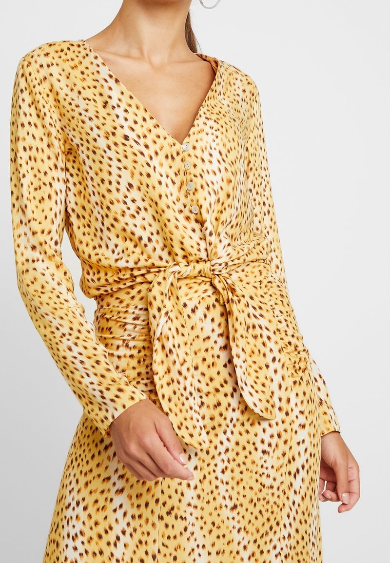 Kvinna som bär en långärmad klänning med gul leopardmönster, framknäppning och knytbart midjebälte, stående mot en enhetlig bakgrund.