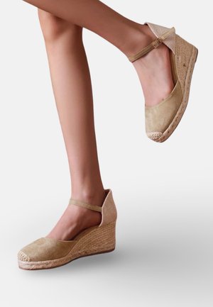 Jambes portant des sandales espadrilles compensées en daim beige avec brides à la cheville sur un fond clair uni.