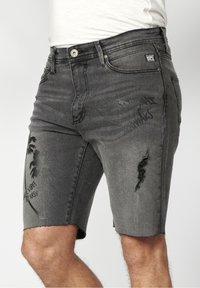 Shorts en denim noir délavé avec détails usés et motif graphique de palmier noir sur la cuisse gauche, portés avec un t-shirt blanc.