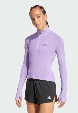 Young woman wearing a fitted lavender zip-up athletic top and black Adidas shorts against a plain background.