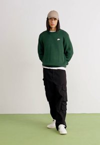 Πράσινο πλεκτό πουλόβερ με μικρό λευκό λογότυπο, μαύρο cargo παντελόνι με τσέπες, λευκά αθλητικά παπούτσια και μπεζ καπέλο, όρθιο σε πράσινο πάτωμα.