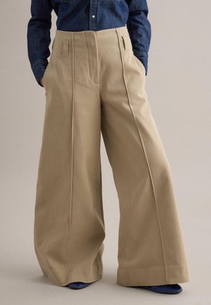 Beige broek met wijde pijpen, voornaad, lussen voor een riem en zijzakken, gedragen met een ingestopt donker denim overhemd en blauwe schoenen.