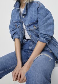 Lichtblauwe denimjas met een losse pasvorm, voorzien van een hoge kraag, knoopsluiting aan de voorkant en twee borstzakken. Draagt bijpassende distressed jeans.