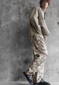 Mladý muž v béžovej košeli a vzorovaných cargo nohaviciach stojí opretý o textúrovanú sivú stenu, pozerá cez plece s rukami v kapsách.