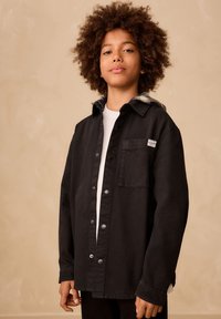 Camicia-giacca nera con cappuccio a quadri grigi, tasca frontale e grandi bottoni; realizzata in tessuto resistente, vestibilità comoda.