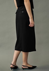Sort knælang nederdel med en tætsiddende talje, der har baglommer og en lille knapdetalje. Båret med sorte slingback sko.