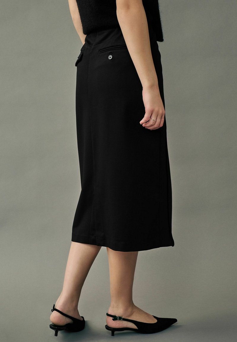 Sort knælang nederdel med en tætsiddende talje, der har baglommer og en lille knapdetalje. Båret med sorte slingback sko.