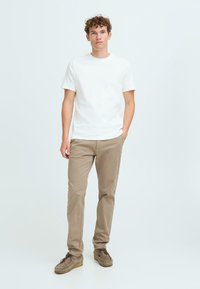White cotton t-shirt, beige pants, and gray suede shoes with brown soles. Model stands with one hand in pocket against a light background.