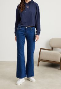 Felpa corta blu navy con logo, abbinata a jeans flare di jeans scuro. La modella indossa sneakers bianche, in piedi su un pavimento neutro.