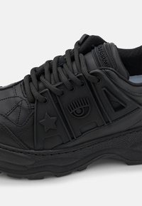 Svart sneaker med texturerad läder, gummiinslag och framträdande ögon- och stjärndesigner. Har kraftiga snören och vadderad krage.