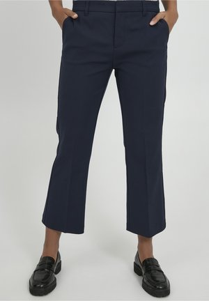 Osoba ubrana w granatowe krótkie spodnie typu flare z bocznymi kieszeniami i czarne masywne loafersy, stojąca na tle jednolitego jasnego tła.