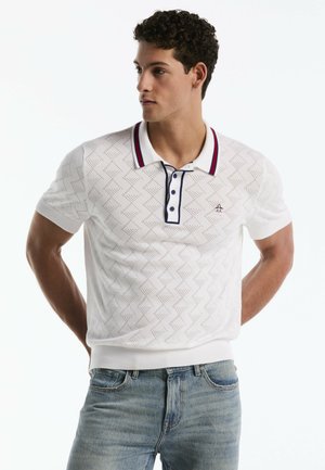 Mann trägt ein weißes kurzärmeliges Poloshirt mit Muster und marineblauen sowie roten Besätzen, kombiniert mit hellblauen Jeans, steht mit den Händen hinter dem Rücken.
