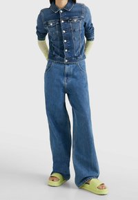 Denim jacket with a collared neckline and four front pockets, paired with wide-leg blue jeans. Footwear features green slide sandals.