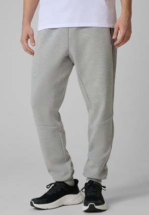 Dolna część ciała osoby noszącej jasnoszare spodnie dresowe i czarne buty sportowe z białymi podeszwami na jednolitym szarym tle.
