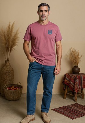 Roodachtige t-shirt met een vierkante grafiek op de linkerborst, gecombineerd met blauwe jeans en beige schoenen, tegen een neutrale achtergrond.