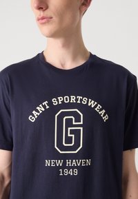 Marineblå bomull t-skjorte med korte ermer, med en kremfarget grafikk av "GANT SPORTSWEAR" og "NEW HAVEN 1949" tekst.