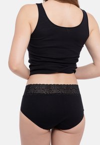 Schwarzes Baumwoll-Tanktop kombiniert mit schwarzen Unterhosen mit Spitzenakzenten. Die Spitzenborte hat ein florales Muster und sorgt für eine kontrastierende Textur.