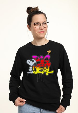 Kobieta w okularach i z kokiem na głowie nosi czarną bluzę z kolorowym graffiti tekstem "Big Deal" i postacią Snoopy'ego.
