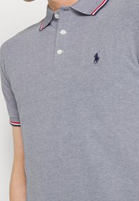 Pikétröja i marinblått och vitt hundtandsmönster, med en tvåknapps knäppning, korta ärmar med röda och vita randiga detaljer, samt logotyp.