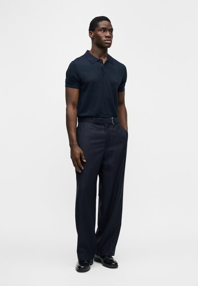 Μπλε σκούρο πόλο με γιακά και ρέλι με κουμπιά, συνδυασμένο με σκούρο παντελόνι με ψιλή γραμμή. Μαύρα παπούτσια ολοκληρώνουν την εμφάνιση.