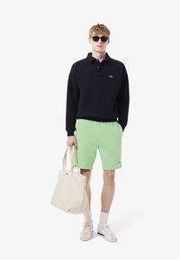 Pull marin avec col, shorts verts, baskets blanches et sac fourre-tout beige. Comprend le logo de la marque sur le pull et les shorts.