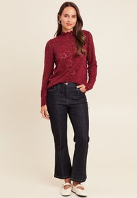 Burgunderfarbenes Spitzenpullover mit hohem Kragen, kombiniert mit dunklen Schlagjeans und weißen Schuhen. Der Pullover hat ein strukturiertes Blumenmuster und lange Ärmel.