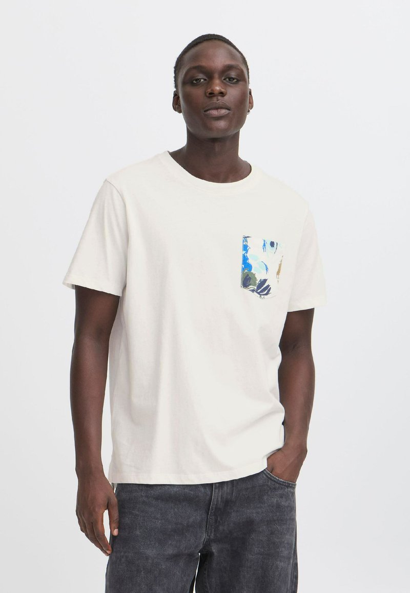 Camiseta de algodón beige claro con cuello redondo y mangas cortas. Presenta un bolsillo impreso con un colorido diseño floral en el lado izquierdo del pecho.