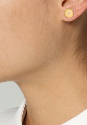 Cou et ligne de la mâchoire d'une personne avec une boucle d'oreille clou ronde en or avec une petite pierre claire au centre.