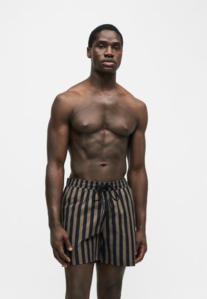 Homme torse nu debout, portant un short de bain à rayures verticales noires et marron avec une taille élastique et un cordon.